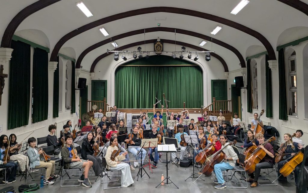 Image shows an orchestra of over 50 students smiling at the camera